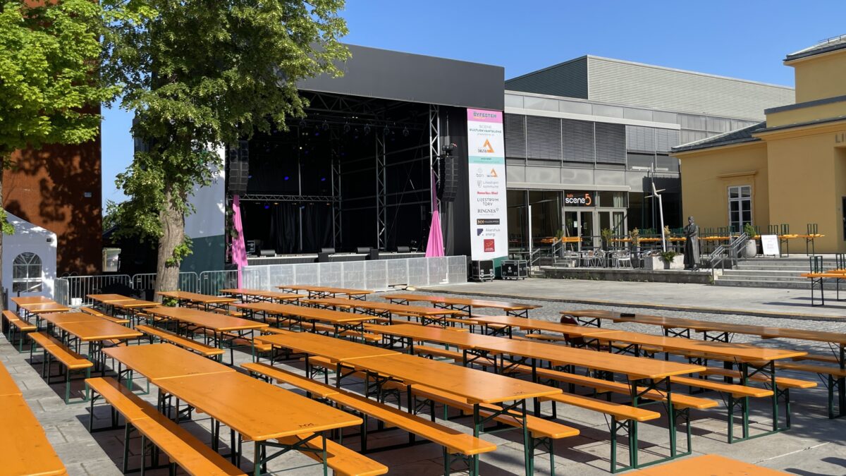 Hallgruppen has set up an outdoor stage with several rows of orange tables and benches, ready for a public event under a clear blue sky.