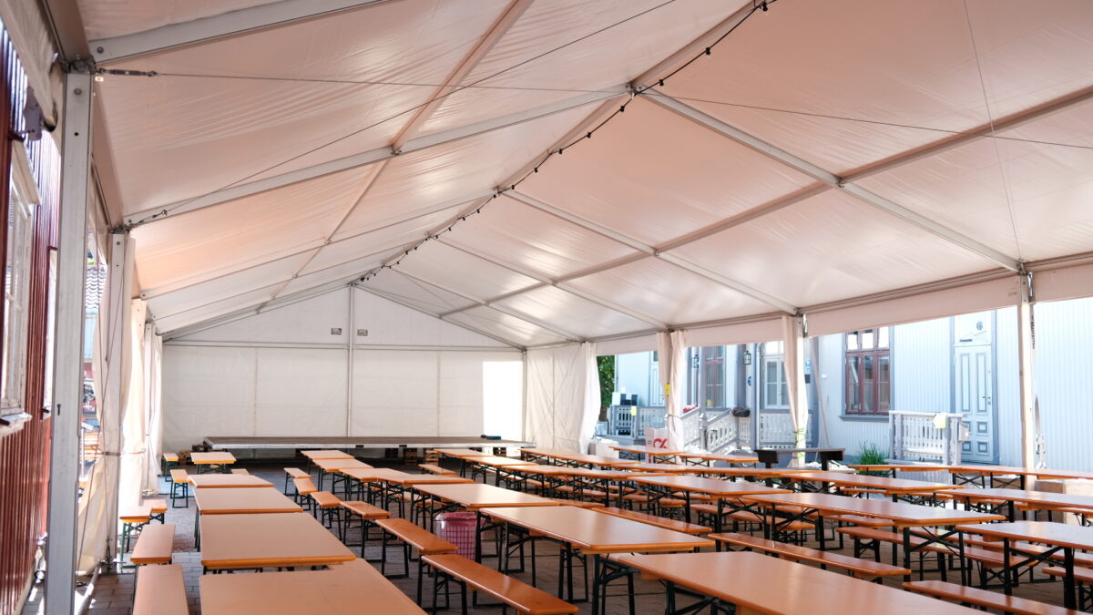 Inside a large white tent, there are several rows of orange tables and benches, arranged for an organized gathering.