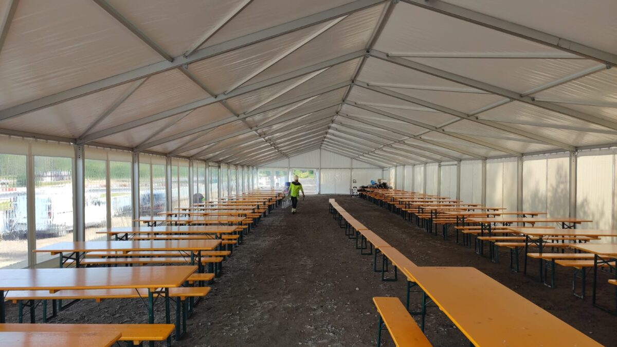 A long white tent with transparent walls, filled with orange tables and benches, set up on a gravel area for an outdoor gathering.